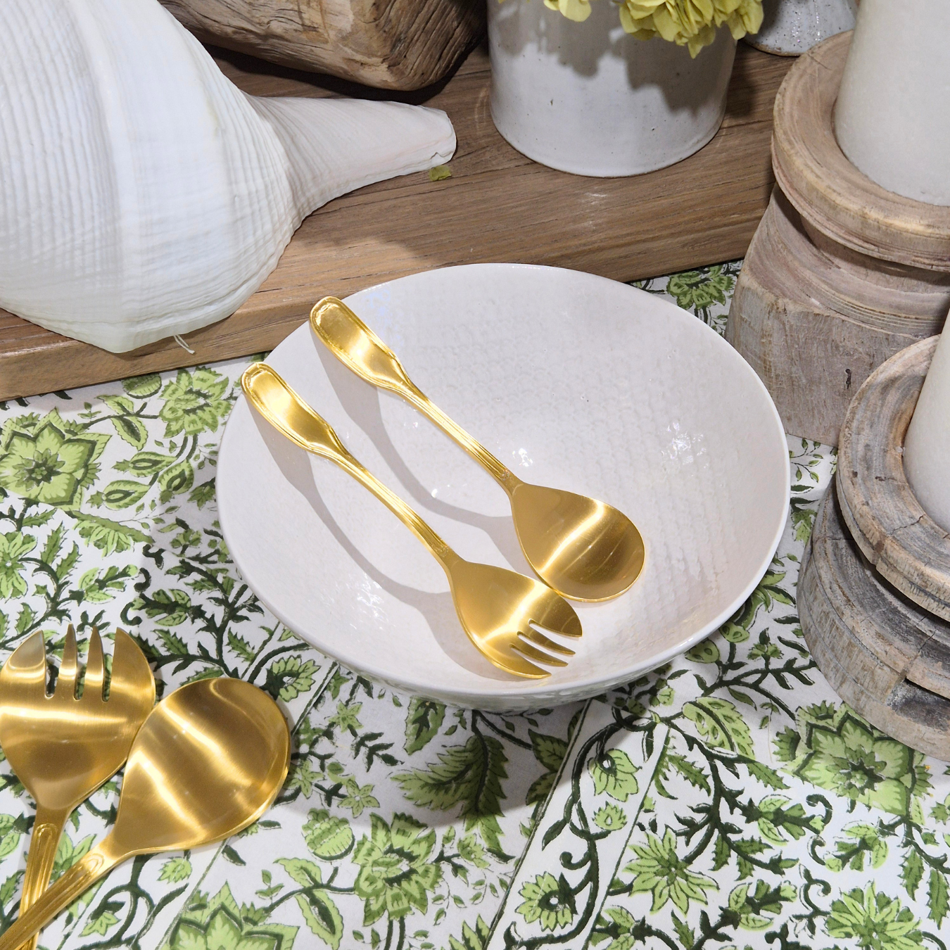 Gold salad servers on a white plate with a floral tablecloth and decorative items in the background