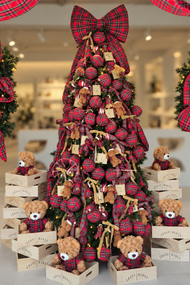 A festive Christmas tree display in a store window, decorated with red and black plaid ornaments, large red and black plaid bows, teddy bears, and wooden crates. Red plaid fabric garlands frame the top of the window display.