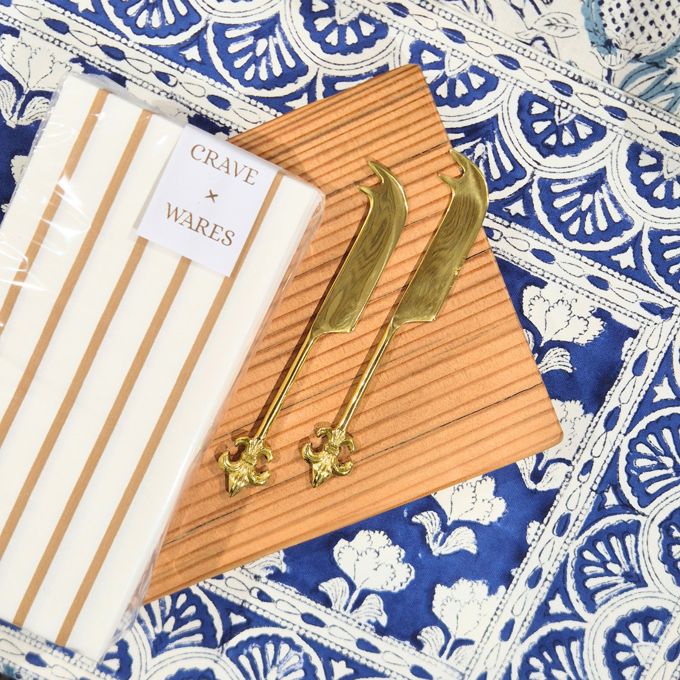 Gold cheese knives on a wooden board with 'Crave' packaging on a patterned fabric background