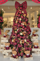 A festive Christmas tree display in a store window, decorated with red and black plaid ornaments, large red and black plaid bows, teddy bears, and wooden crates. Red plaid fabric garlands frame the top of the window display.
